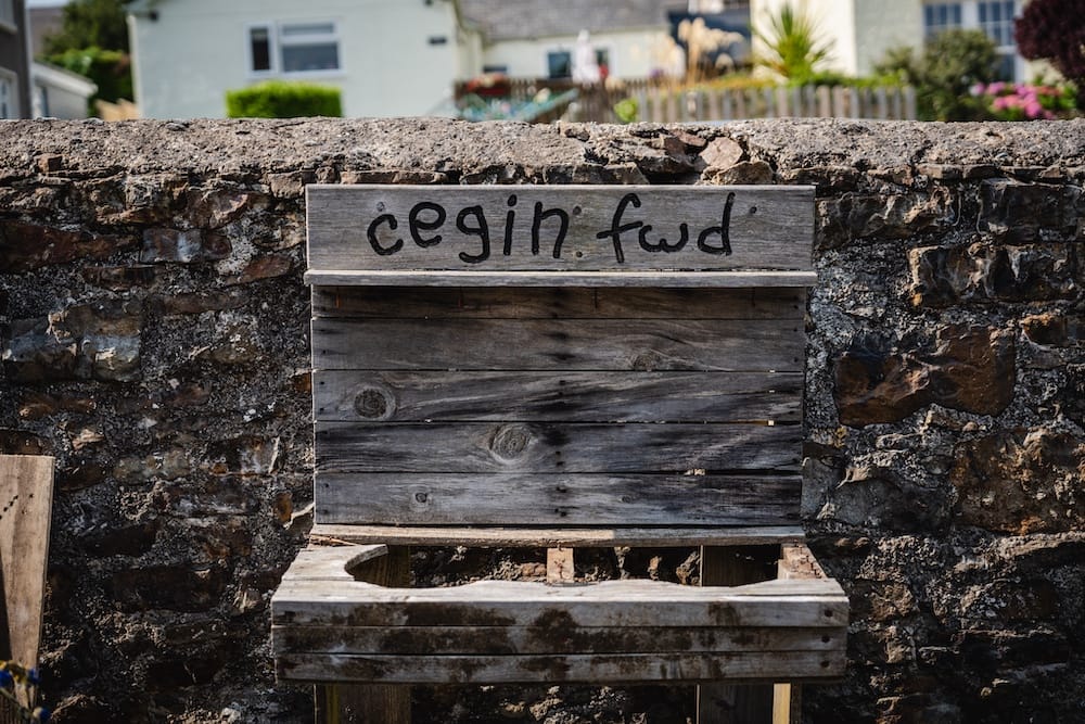 Children's mud kitchen infront of a stone wall with 'cegin fwd' written on a wooden plank
